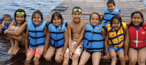 Neuf enfants, âgés d'environ 7 à 11 ans, portent des gilets de sauvetage et des maillots de bain et sourient pour une photo en plein air, assis sur un quai, au bord d'un lac.