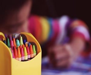 Un enfant desinne derrière une boite de crayons colorés